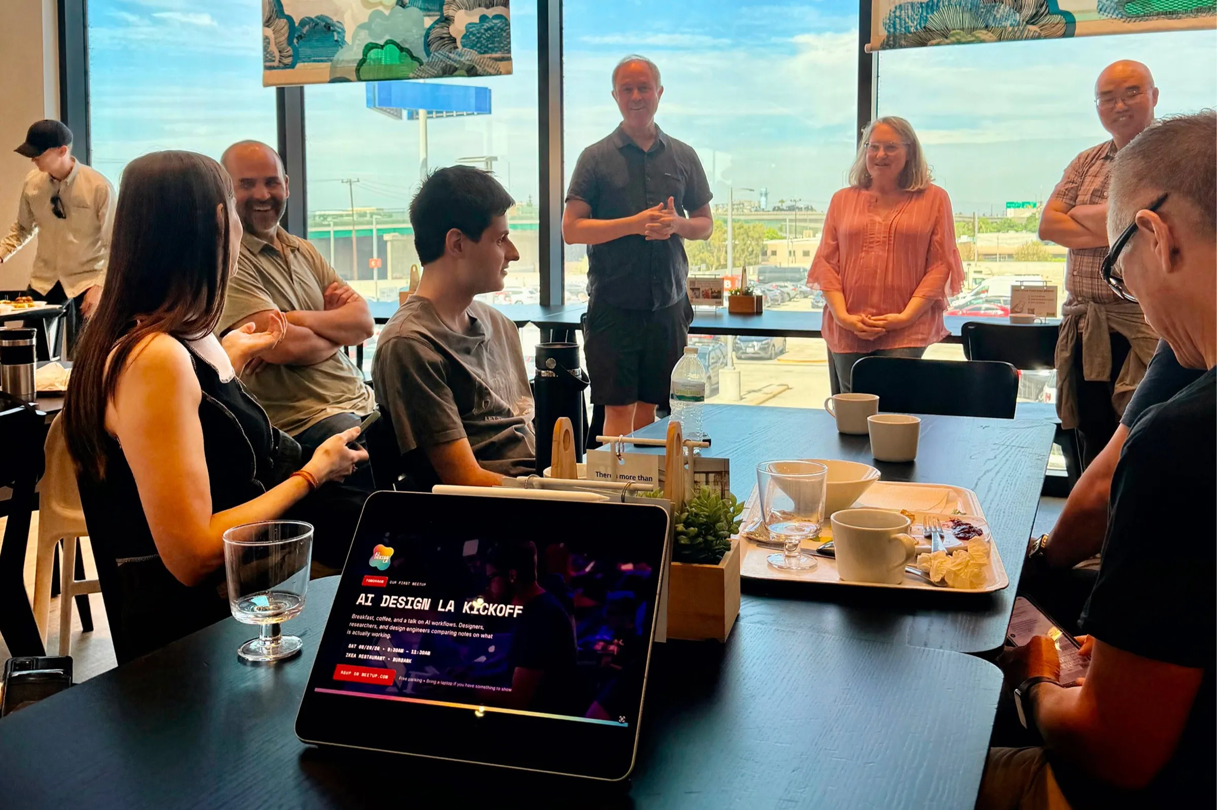 Members at an AI Design LA meetup gathered around a table in a Los Angeles cafe, with the community's kickoff event page open on a tablet.
